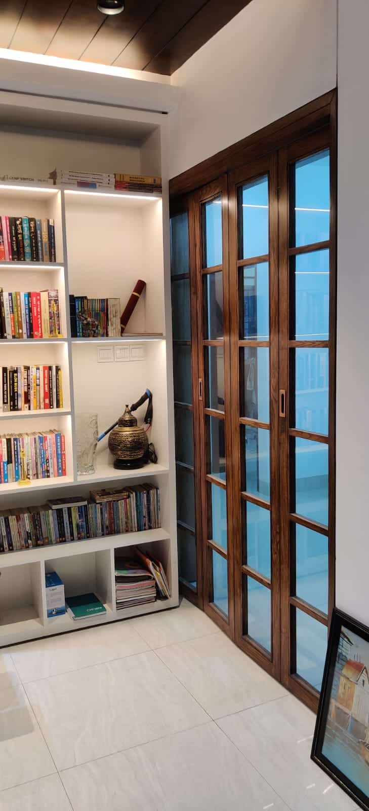 Tall bookshelf with books and decor beside a wooden-frame glass folding door - residence interior design in Rampura, Dhaka
